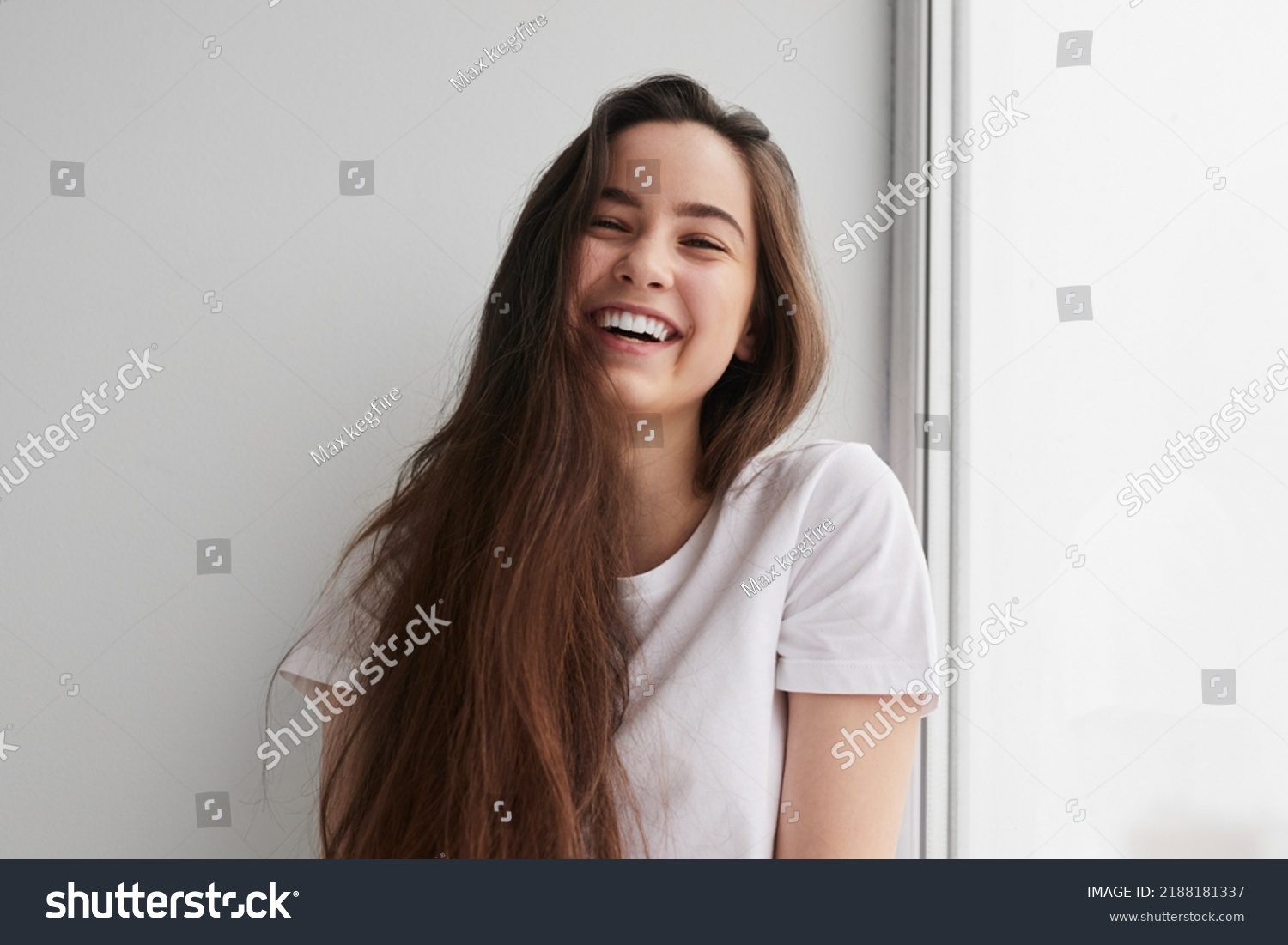 Delighted young lady with long dark hair in t shirt smiling happily and looking at camera, while sitting near window and leaning on white wall #2188181337