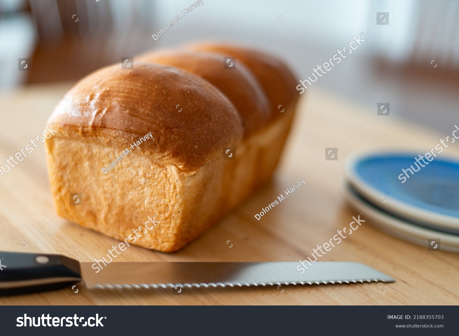 A single loaf of fresh white crusty bread on a wooden cutting board on a kitchen table. The warm crisp bun has melted butter over the crisp loaf buns. Side plates and a knife are next to the loaf.  #2188355703