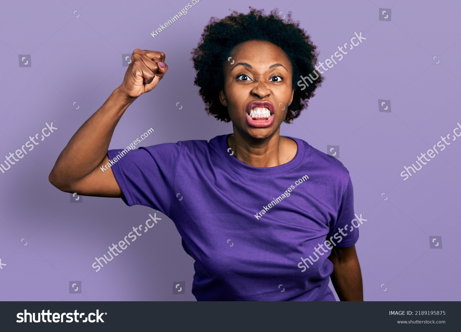 African american woman with afro hair wearing casual purple t shirt angry and mad raising fist frustrated and furious while shouting with anger. rage and aggressive concept.  #2189195875