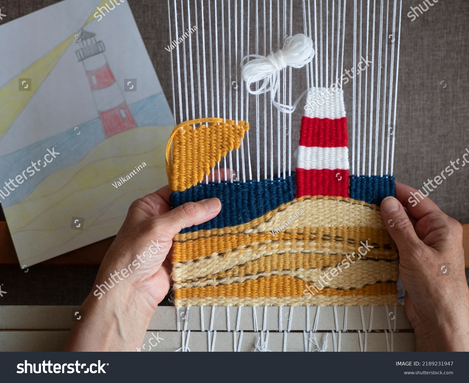 Weaver are making a tapestry with lighthouse following the sketch. Tapestry weaving, selective focus. Hands of artisan at work, close up #2189231947