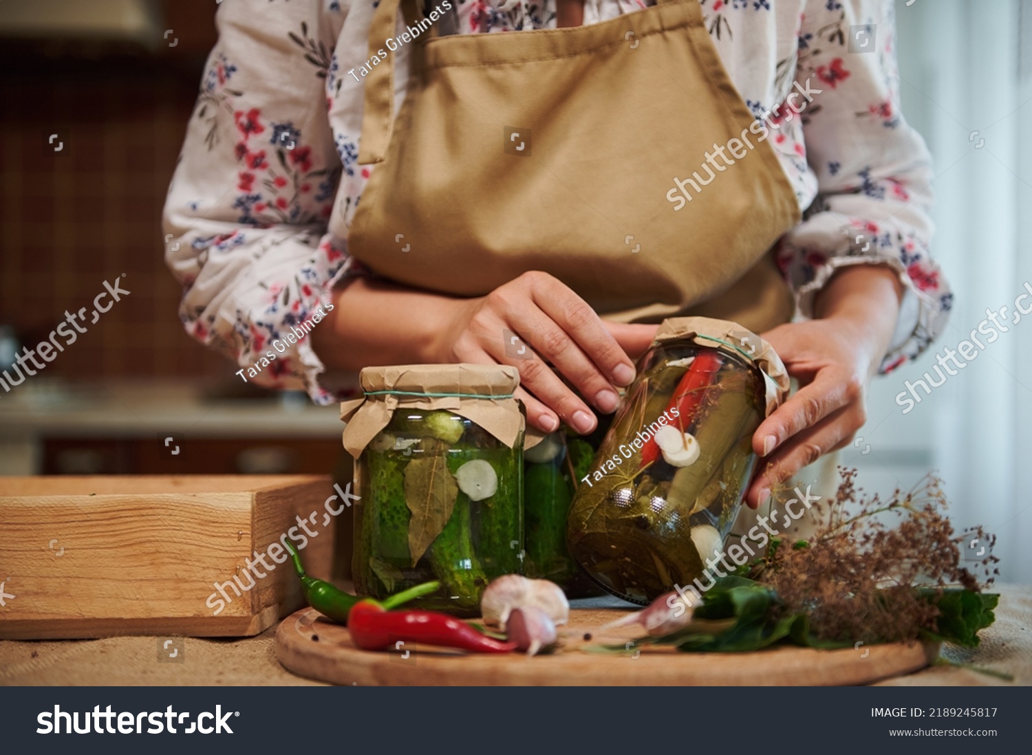 Close-up of homemade pickled canned cucumbers and chili peppers in sterilized glass jars in the hands of a young woman housewife. Fresh fragrant ingredients and culinary herbs on a kitchen table #2189245817