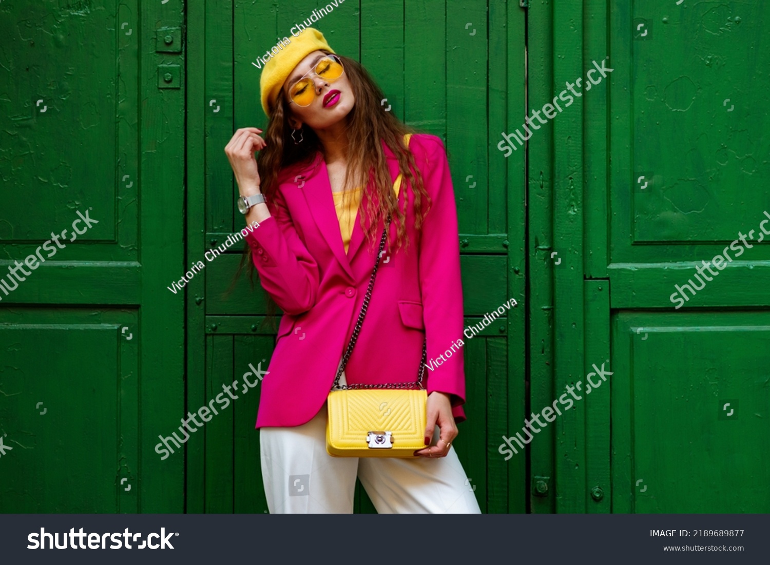Fashionable confident woman wearing trendy outfit with yellow sunglasses, beret, wrist watch, shoulder bag, pink fuchsia color blazer, posing near green door. Copy, empty space for text #2189689877