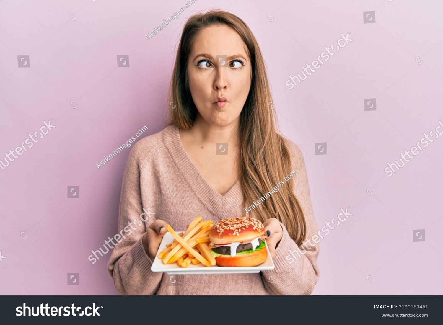 Young blonde woman eating a tasty classic burger with fries making fish face with mouth and squinting eyes, crazy and comical.  #2190160461