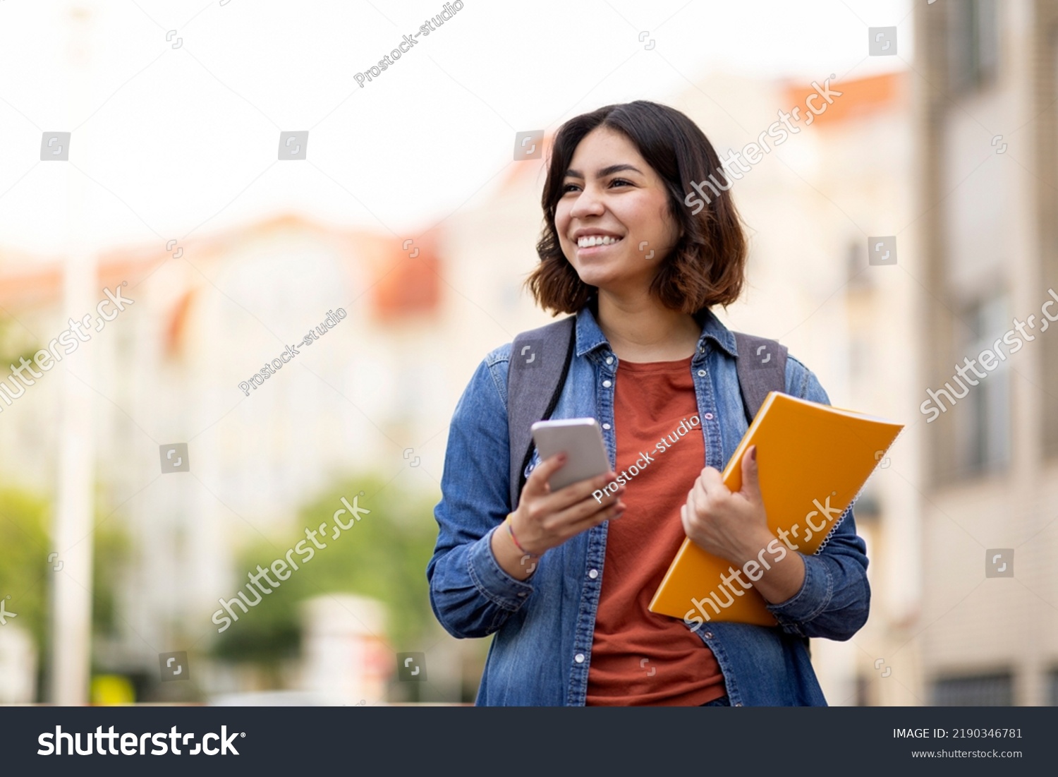 Cheerful Arab Female Student With Smartphone And Workbooks Standing Outdoors, Happy Young Middle Eastern Woman Walking In City After College Classes, Looking Away And Smiling, Copy Space #2190346781
