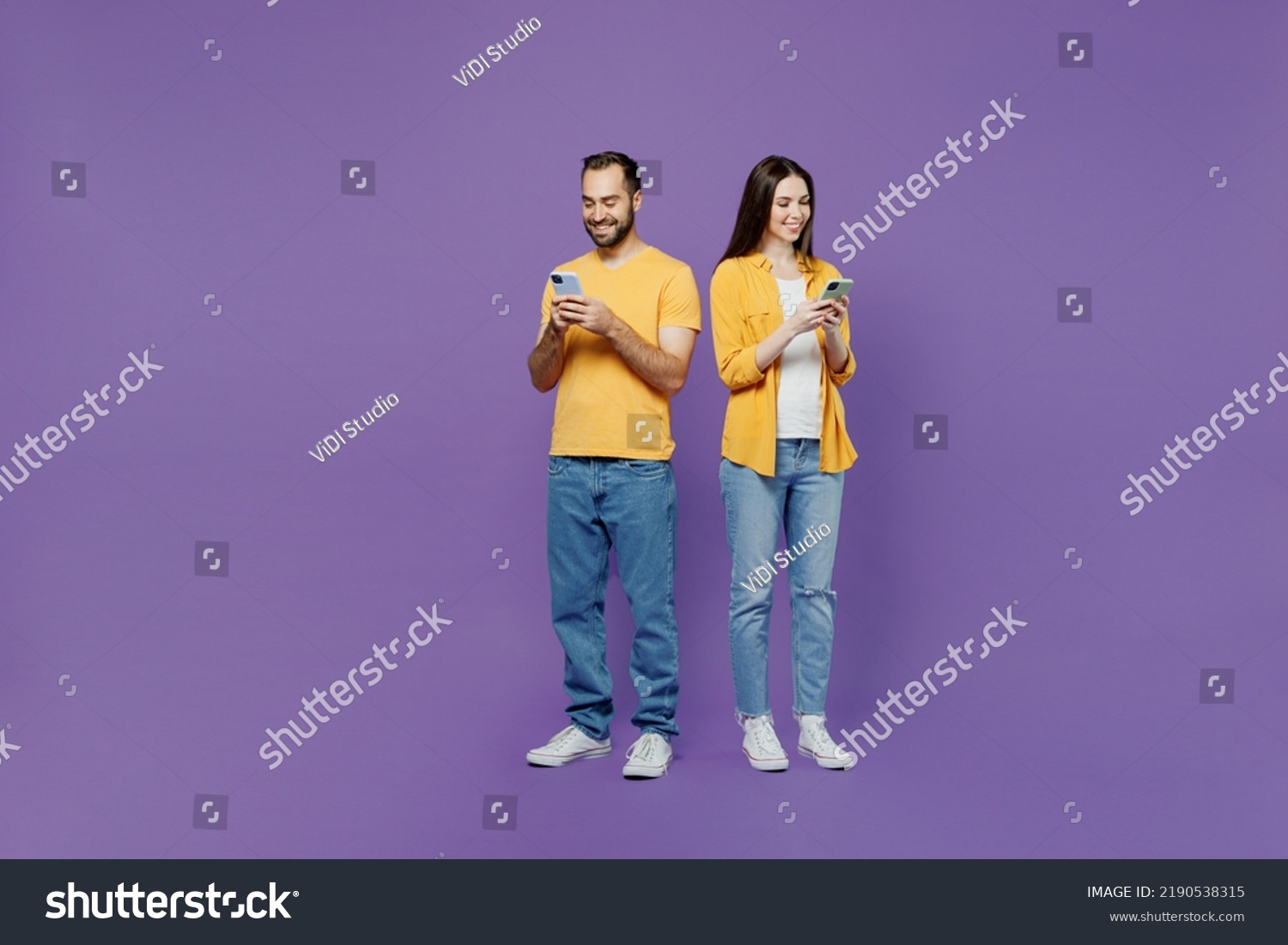 Full body young couple two friends family man woman together in yellow casual clothes hold in hand use mobile cell phone browsing internet chatting online isolated on plain violet background studio #2190538315