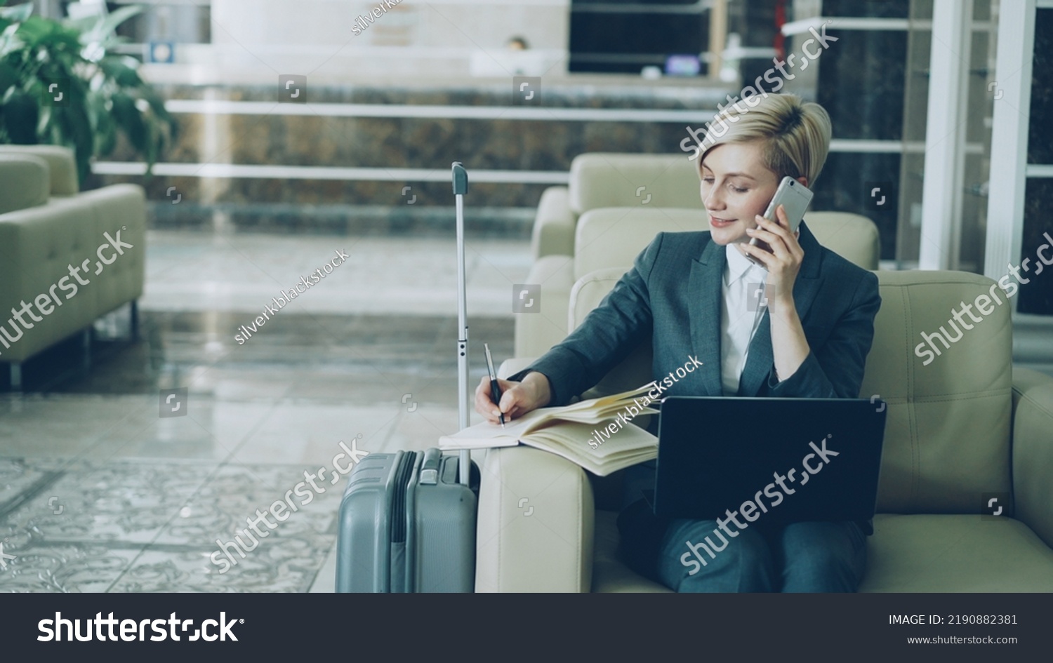 Pan shot of blonde busy businesswoman sitting in armchair in hotel lobby talking mobile phone, writing in notepad and using laptop computer smiling. Business, travel and people concept #2190882381