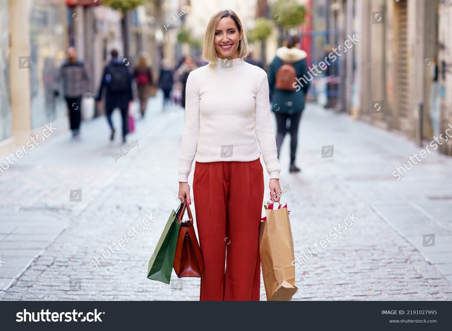 Satisfied female shopper in white turtleneck and pants standing with shopping packages and handbag on paved street and smiling at camera #2191027995