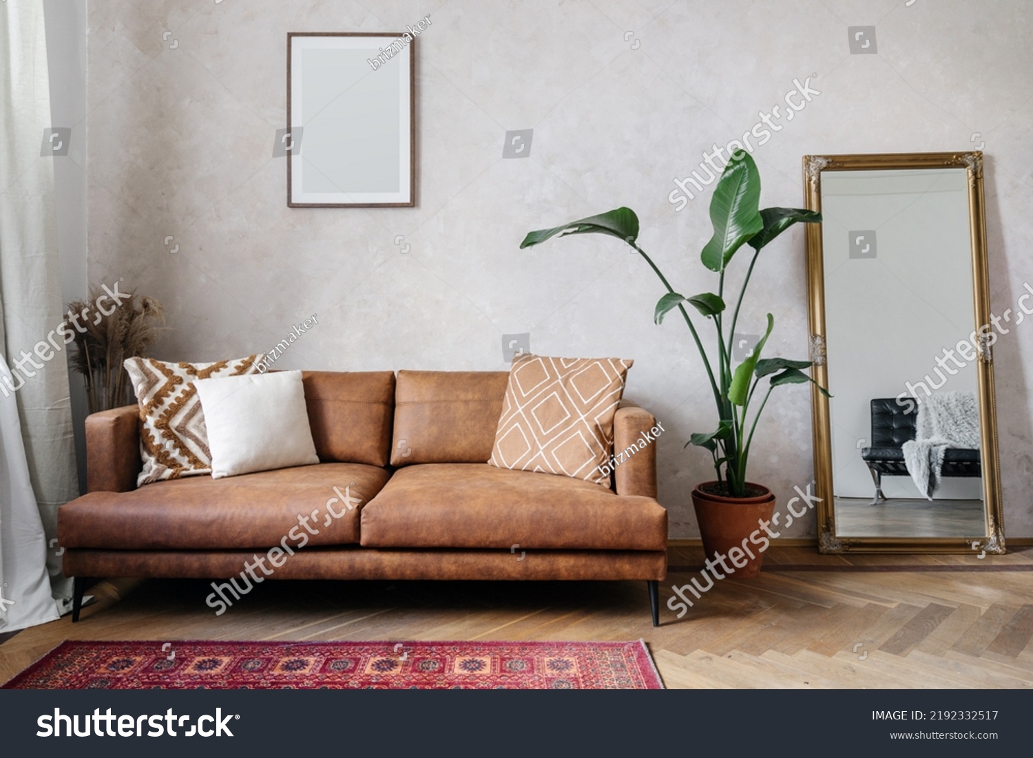 Modern design of living room with brown eco leather couch, soft cushions, mirror with golden frame, copy space picture frame on wall and houseplant in pot #2192332517