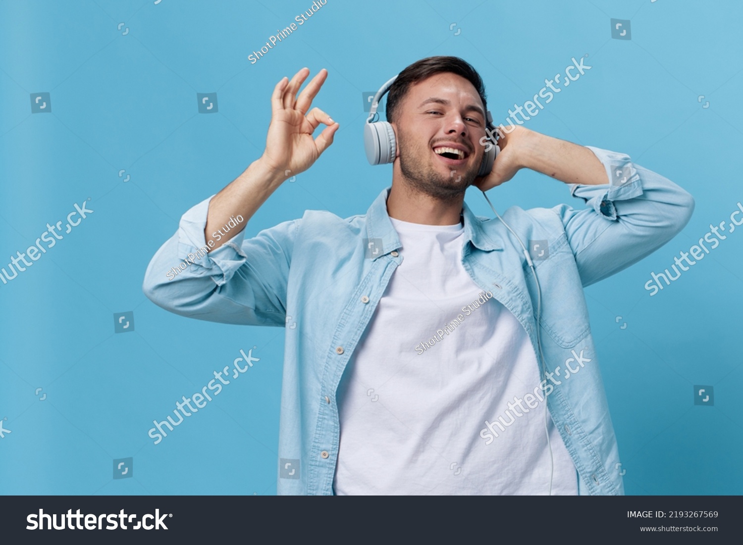 Happy attractive tanned handsome man in casual basic t-shirt headphones listen cool sound show gesture Okay posing isolated on blue studio background. Copy space Banner Mockup. Music concept #2193267569