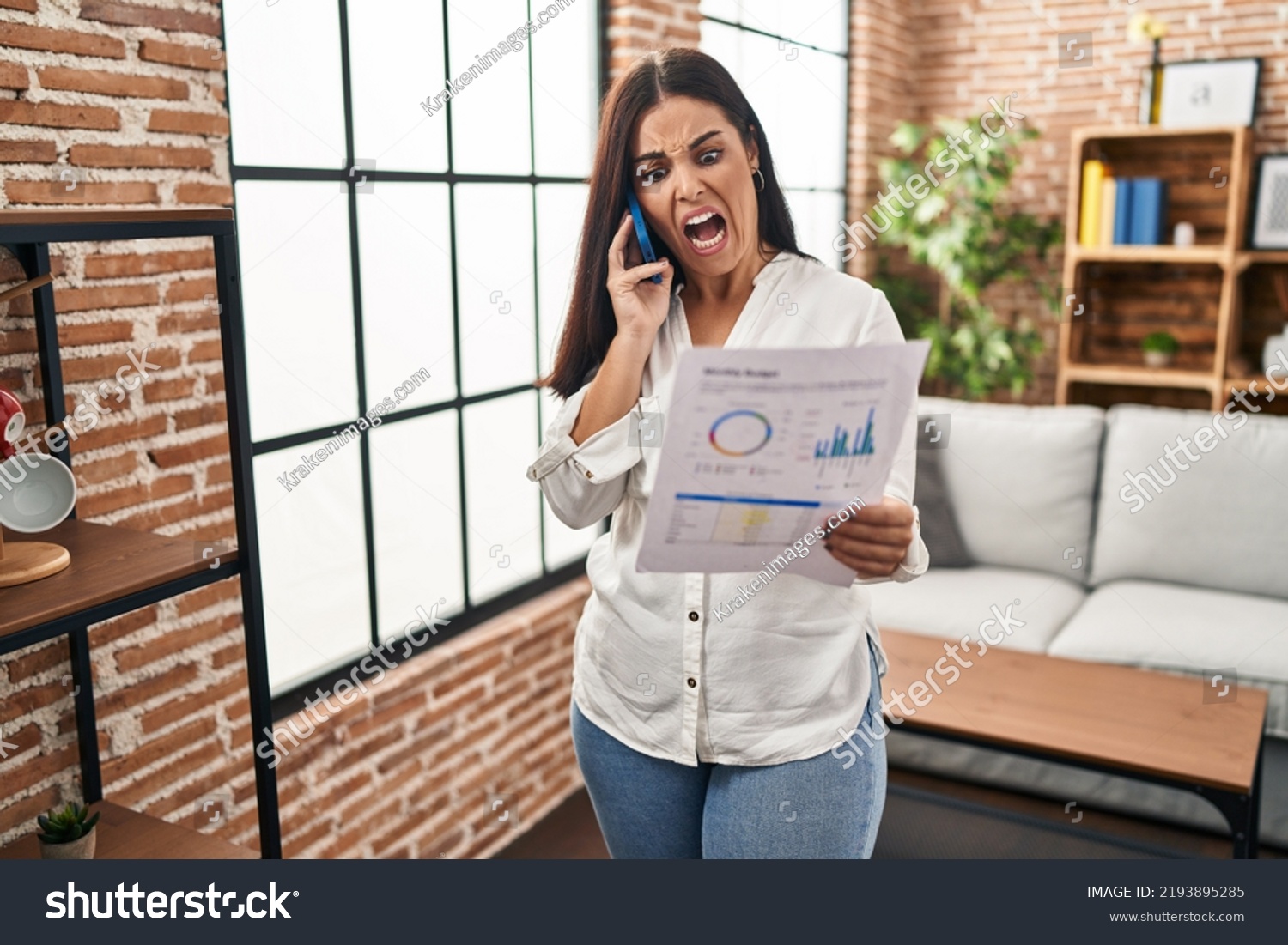 Young hispanic woman speaking on the phone about bills angry and mad screaming frustrated and furious, shouting with anger. rage and aggressive concept.  #2193895285