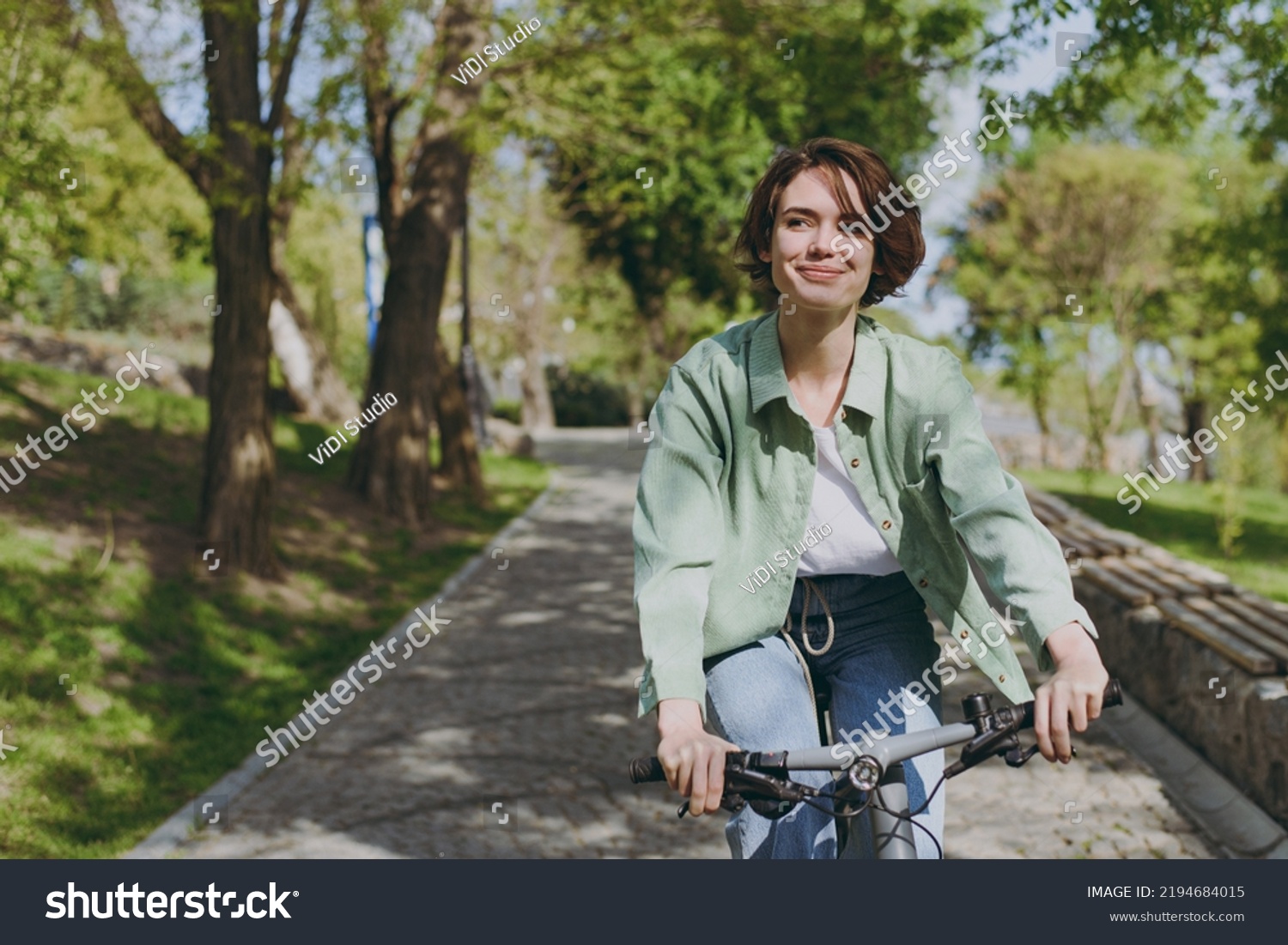 Young pensive dreamful happy woman 20s wearing casual green jacket jeans riding bicycle bike on sidewalk in city spring park outdoors, look aside. People active urban healthy lifestyle cycling concept #2194684015
