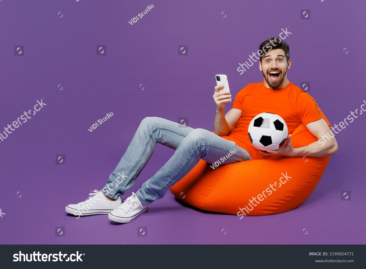 Full size young fan man he wear orange t-shirt cheer up support football sport team hold soccer ball mobile cell phone watch tv live stream sit in bag chair isolated on plain dark purple background #2195824771