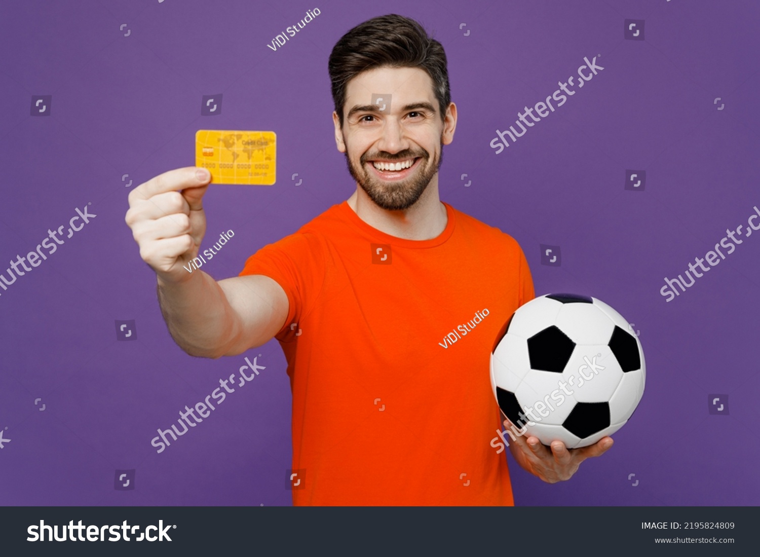 Young happy fan smiling man he wear orange t-shirt cheer up support football sport team hold in hand soccer ball mock up of credit bank card watch tv live stream isolated on plain purple background #2195824809