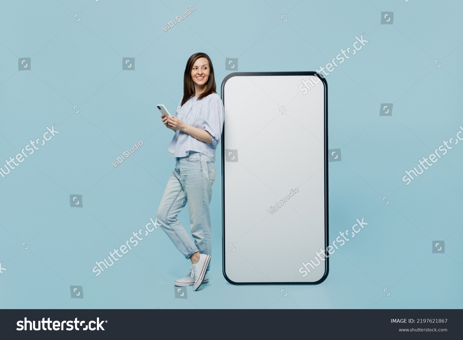 Full size young happy woman she 20s in casual blouse big huge blank screen mobile cell phone with workspace copy space mockup area hold smartphone isolated on pastel plain light blue background studio #2197621867