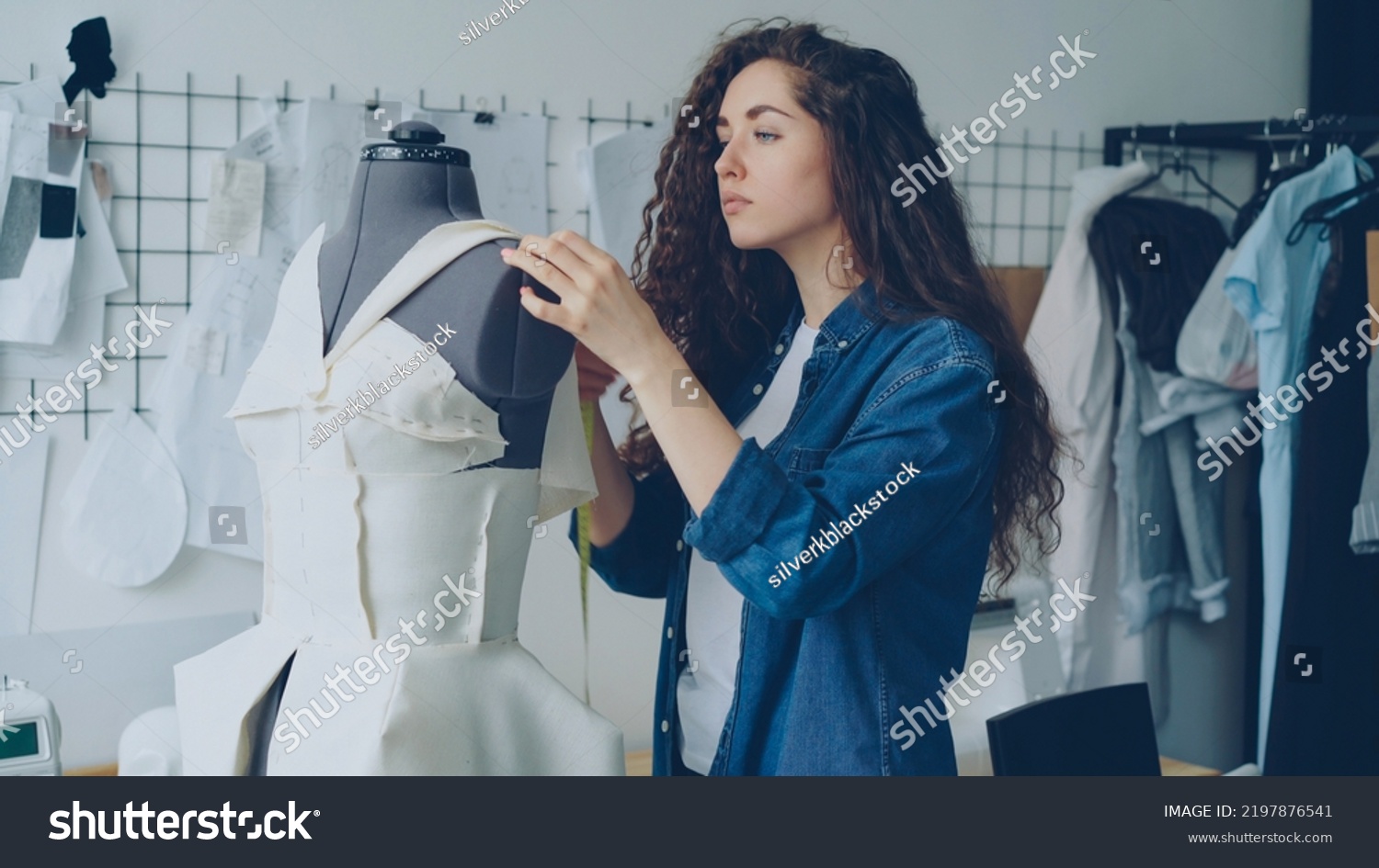 Young female tailor is adjusting clothes on tailoring dummy with sewing pins and measuring with measure-tape. Women's garments, sketches on wall, tailoring items and tools are visible. #2197876541