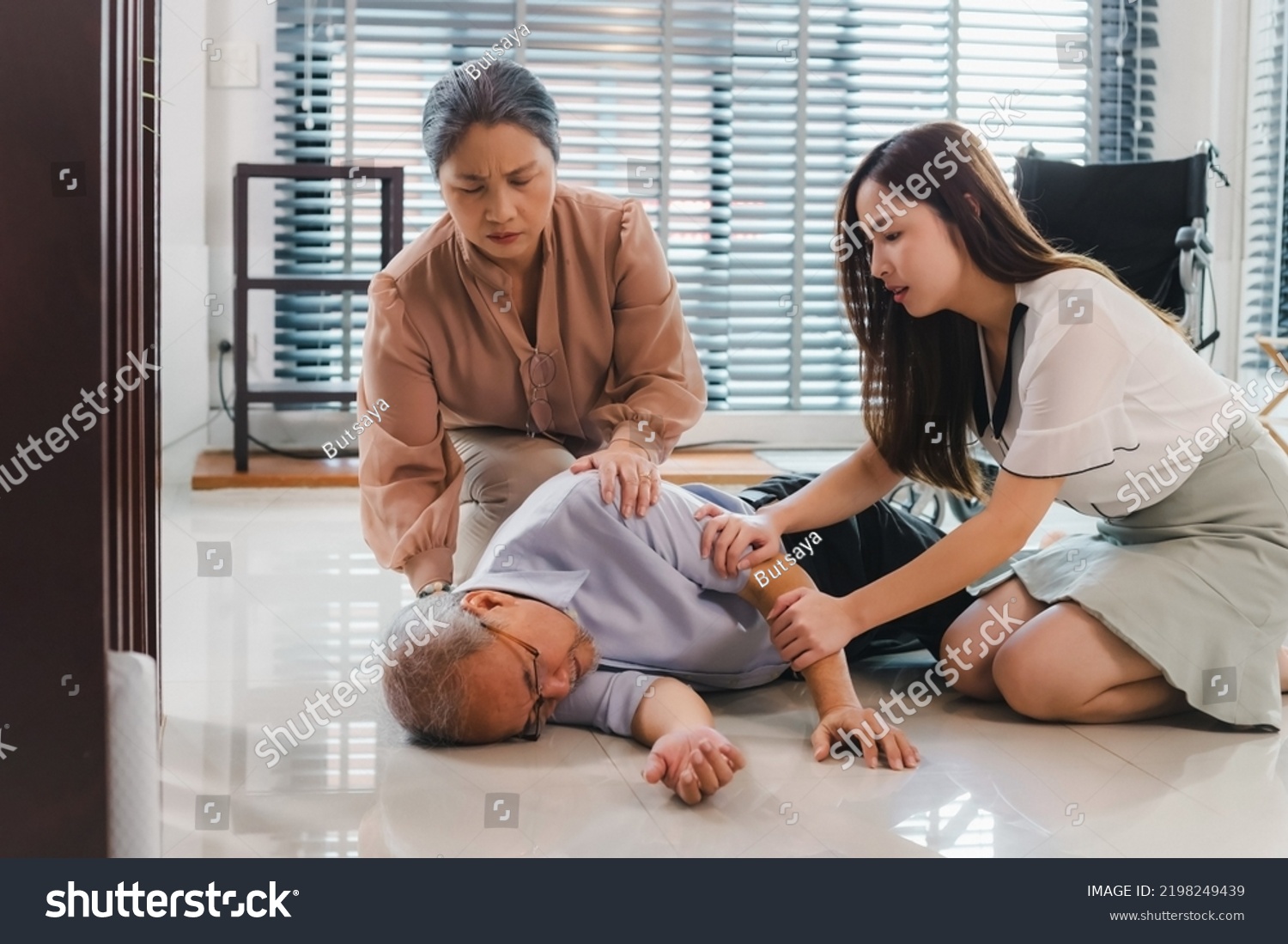 nervous Asian woman granddaughter and wife shaking her grandfather trying to awake him. the old man loses conscious falling on the floor in living room at home. senior man fallen from wheelchair. #2198249439