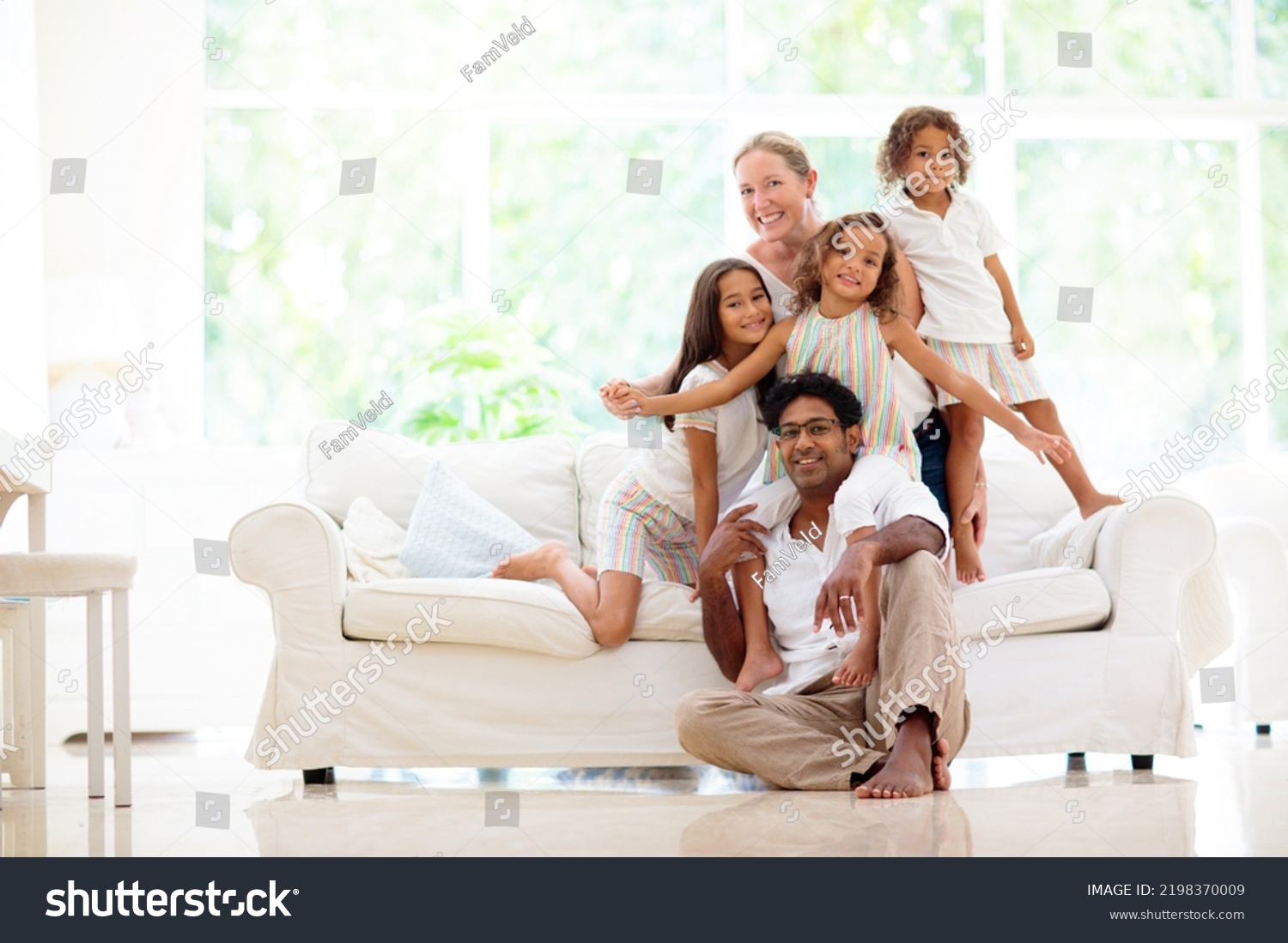 Happy family with kids at home. Parents and children sitting on white couch in sunny living room. Mother, father, son and daughter play and laugh. Young beautiful interracial couple in new house. #2198370009