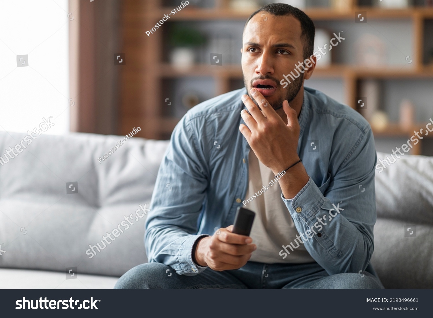 Shocked Black Man With Remote Controller In Hand Sitting On Couch At Home, Confused Young African American Guy Watching Tv Show In Living Room, Emotionally Reacting To Shocking Content, Copy Space #2198496661