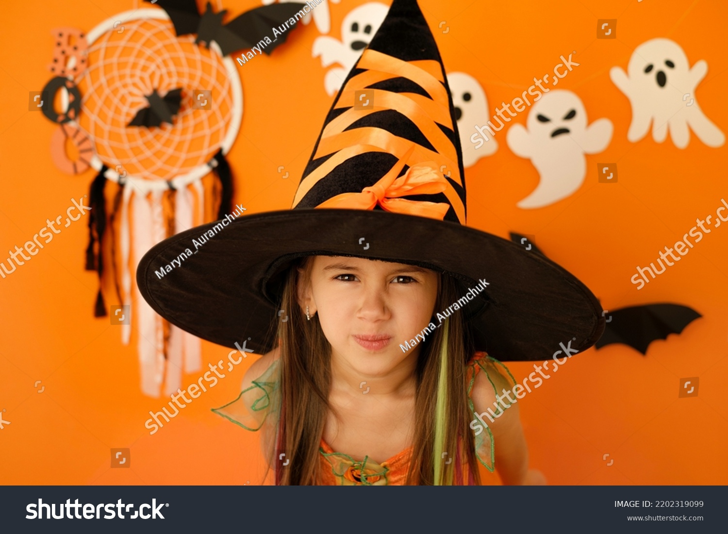 An angry dissatisfied girl in the guise of a witch looks into the camera making a face. Halloween decor with ghosts on an orange studio background #2202319099