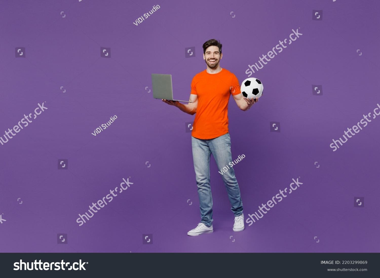 Full body young fan man wear orange t-shirt cheer up support football sport team hold soccer ball watch tv live stream 2024 use betting on laptop pc computer isolated on plain dark purple background #2203299869