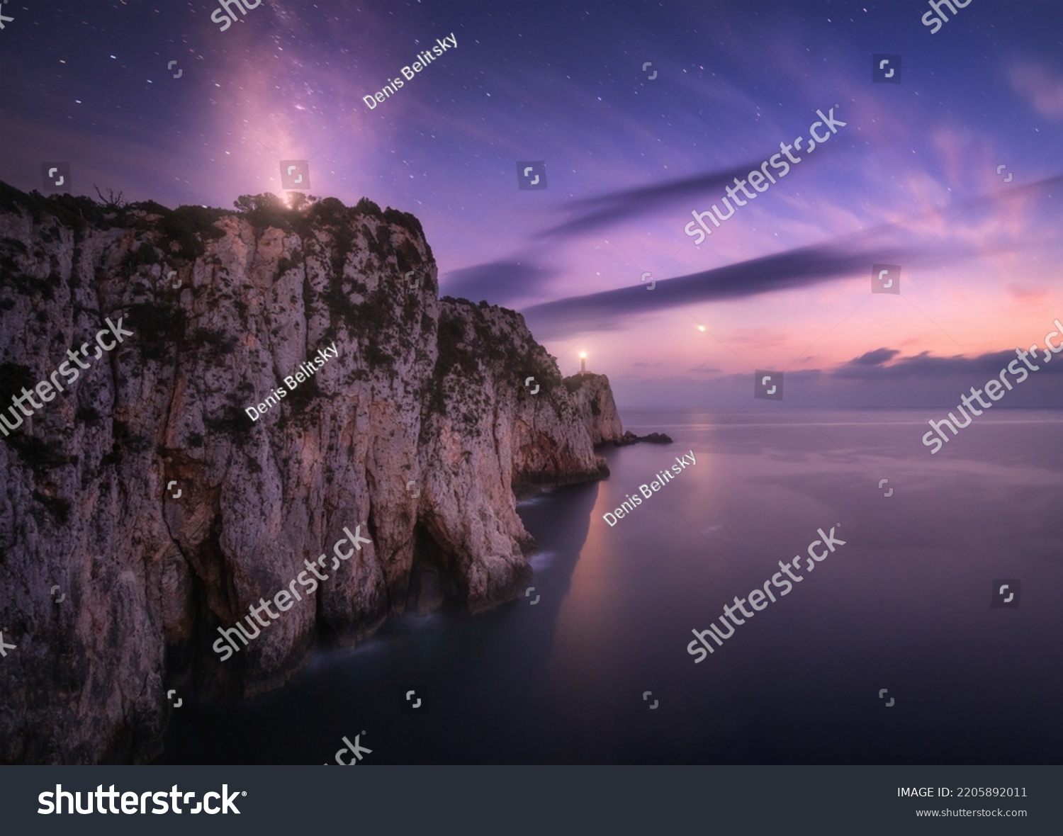 Lighthouse on the mountain peak at starry night in summer. Beautiful cliffs, rocky sea coast, bright stars, milky way and violet sky with clouds at dusk. Lighthouse of Cape Lefkada, Greece. Landscape #2205892011