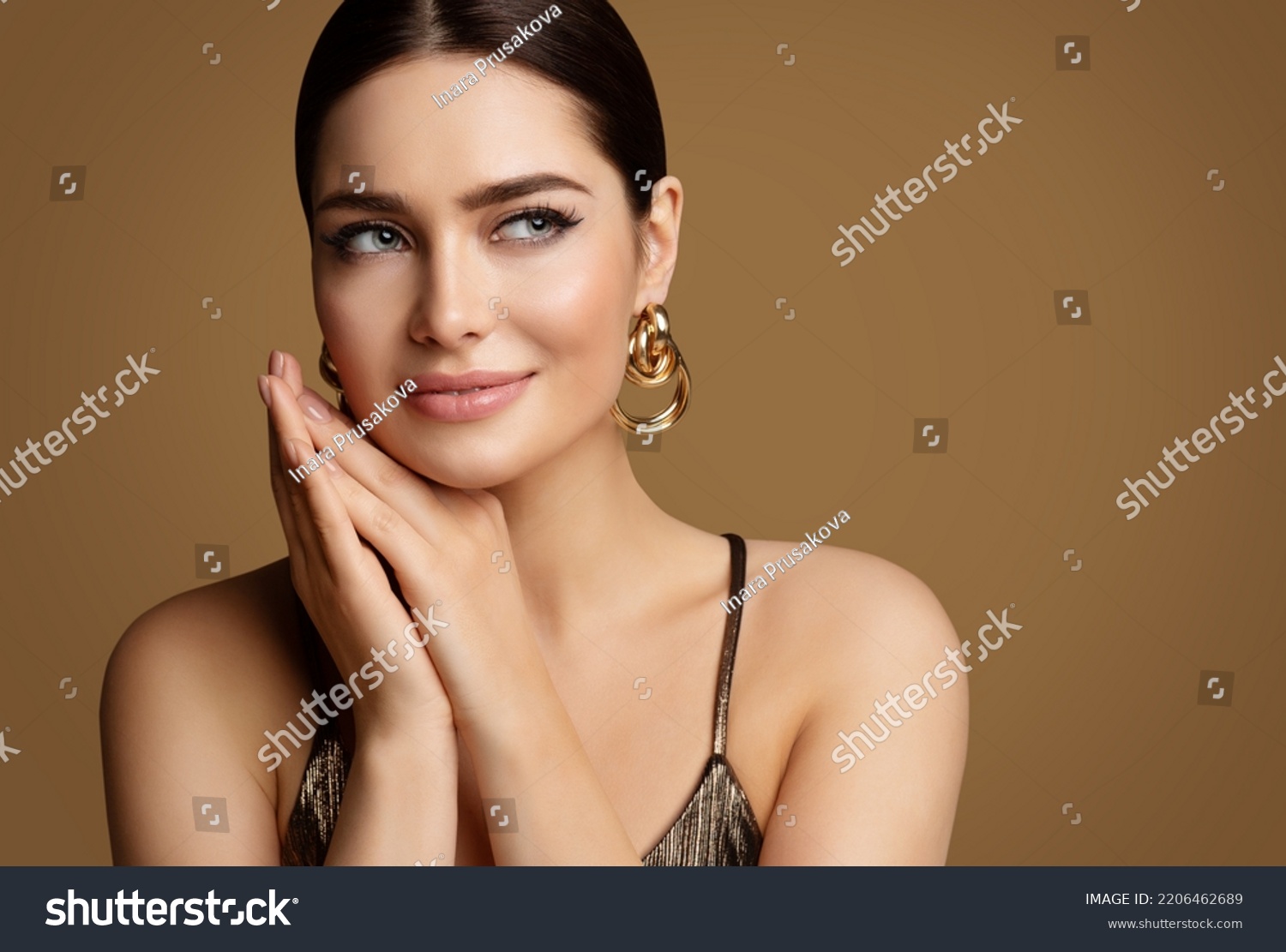Woman Beauty with Smooth Skin Make up and Golden Jewelry. Beautiful Girl with Perfect Lips and Eye Makeup holding Hands under Chin. Elegant Model Portrait with Gold Earring smiling #2206462689