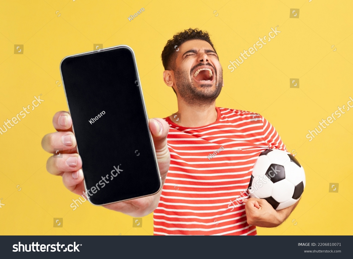 Extremely happy rejoicing bearded man in red T-shirt showing cell phone with blank display and holding soccer ball, celebrating his winning, betting. Indoor studio shot isolated on yellow background. #2206810071