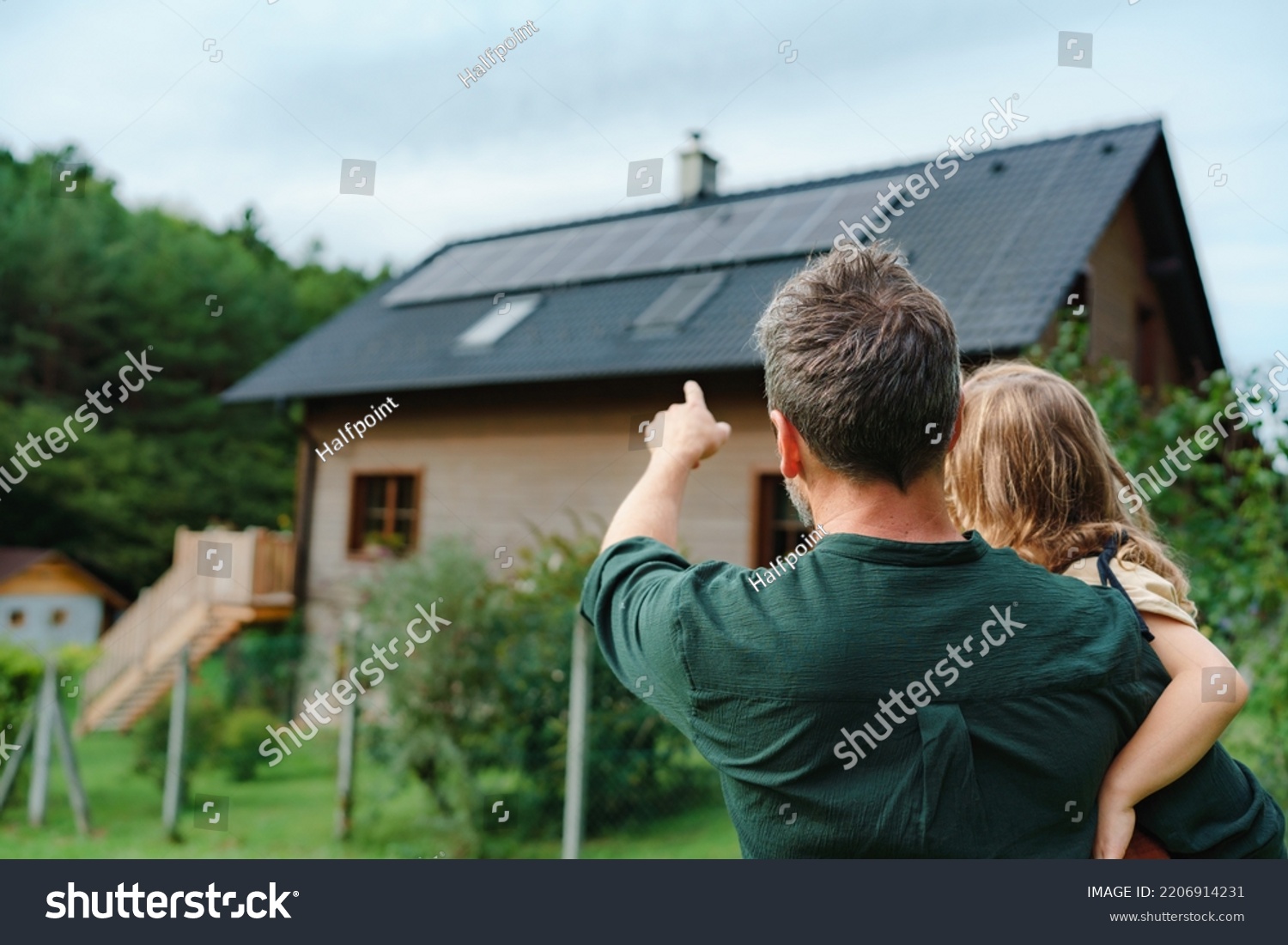 Rear view of dad holding her little girl in arms and showing at their house with installed solar panels. Alternative energy, saving resources and sustainable lifestyle concept. #2206914231