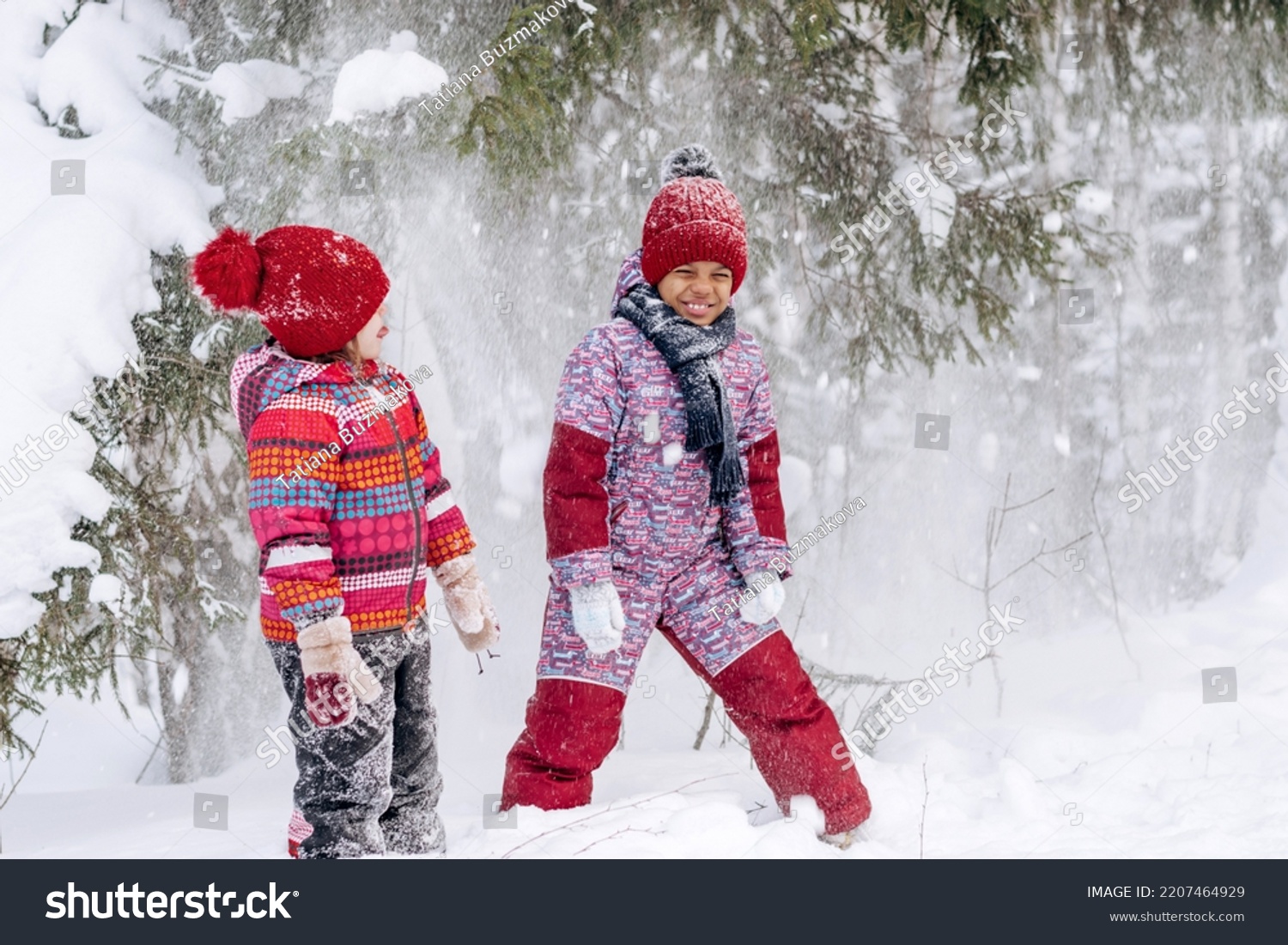 Happy Caucasian and African-American girls are walking in a winter park, shaking snow from snow-covered trees.Winter fun,active lifestyle concept. #2207464929