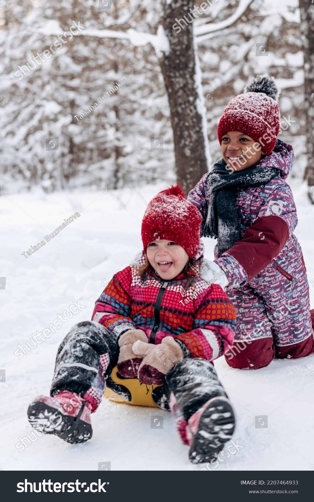 Happy Caucasian and African-American girls ride a saucer in the winter park.Beautiful trees are covered with white snow.Winter fun,active lifestyle concept. #2207464933