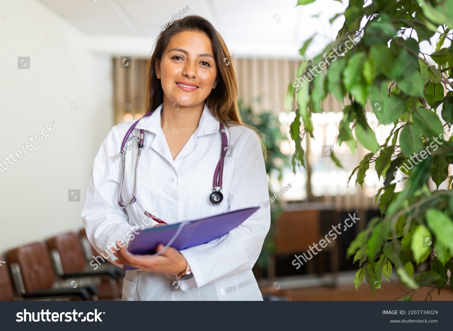 Portrait of young latin american female doctor wearing white coat standing in clinic office, filling out medical form at clipboard. #2207734029