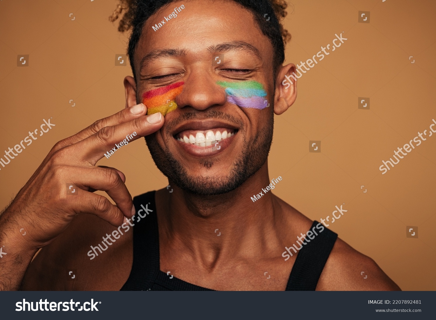 Cheerful African American gay smiling with closed eyes and painting rainbow on cheeks during LGBT pride event against brown background #2207892481