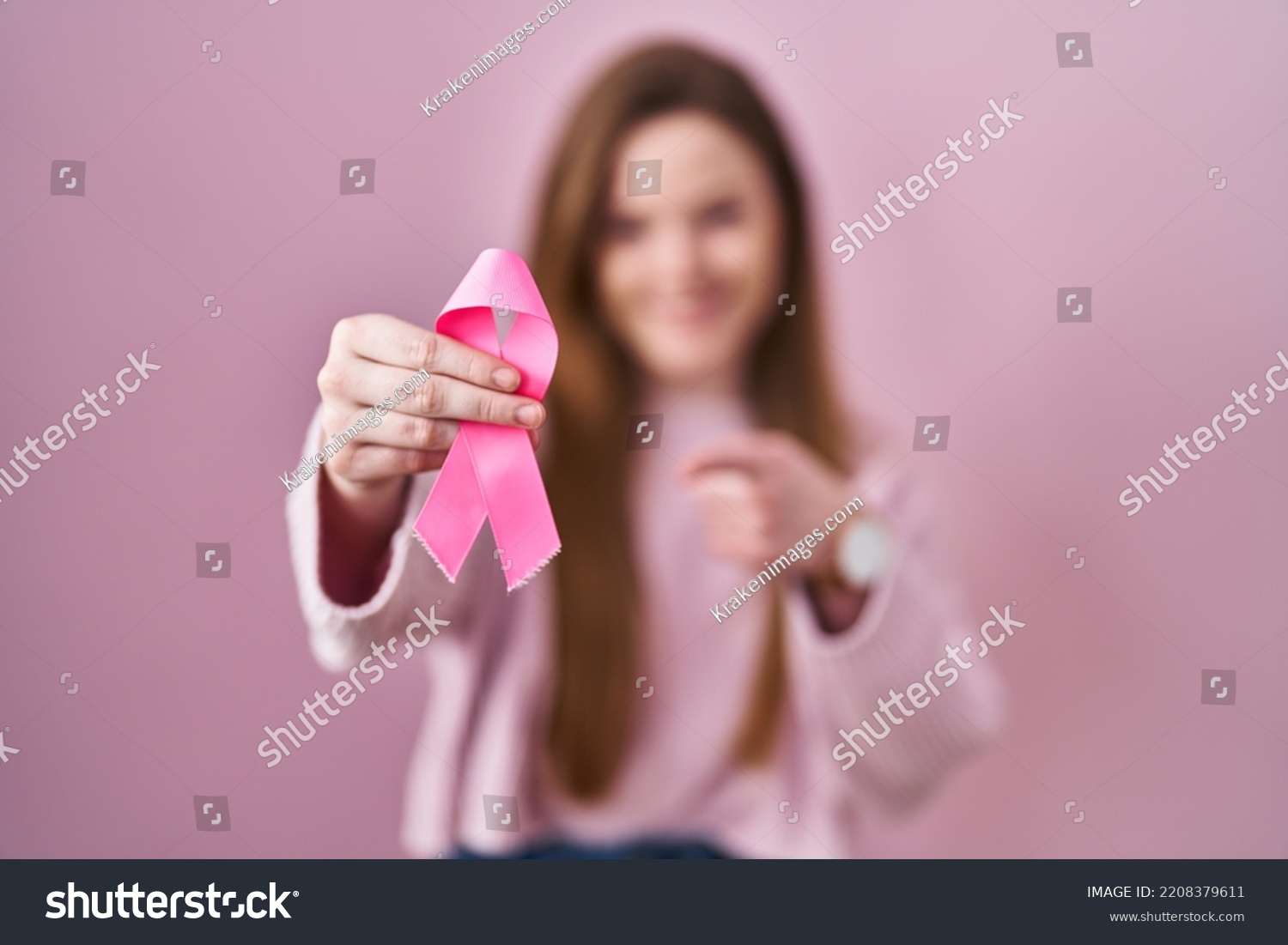 Young caucasian woman holding pink cancer ribbon smiling happy pointing with hand and finger  #2208379611