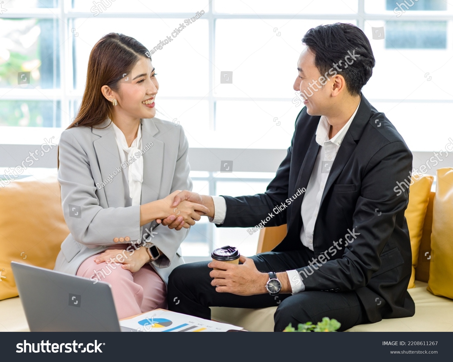 Millennial Asian professional successful businessman in formal suit sitting on sofa smiling holding disposable hot coffee cup shaking hands greeting with businesswoman customer in office meeting room. #2208611267
