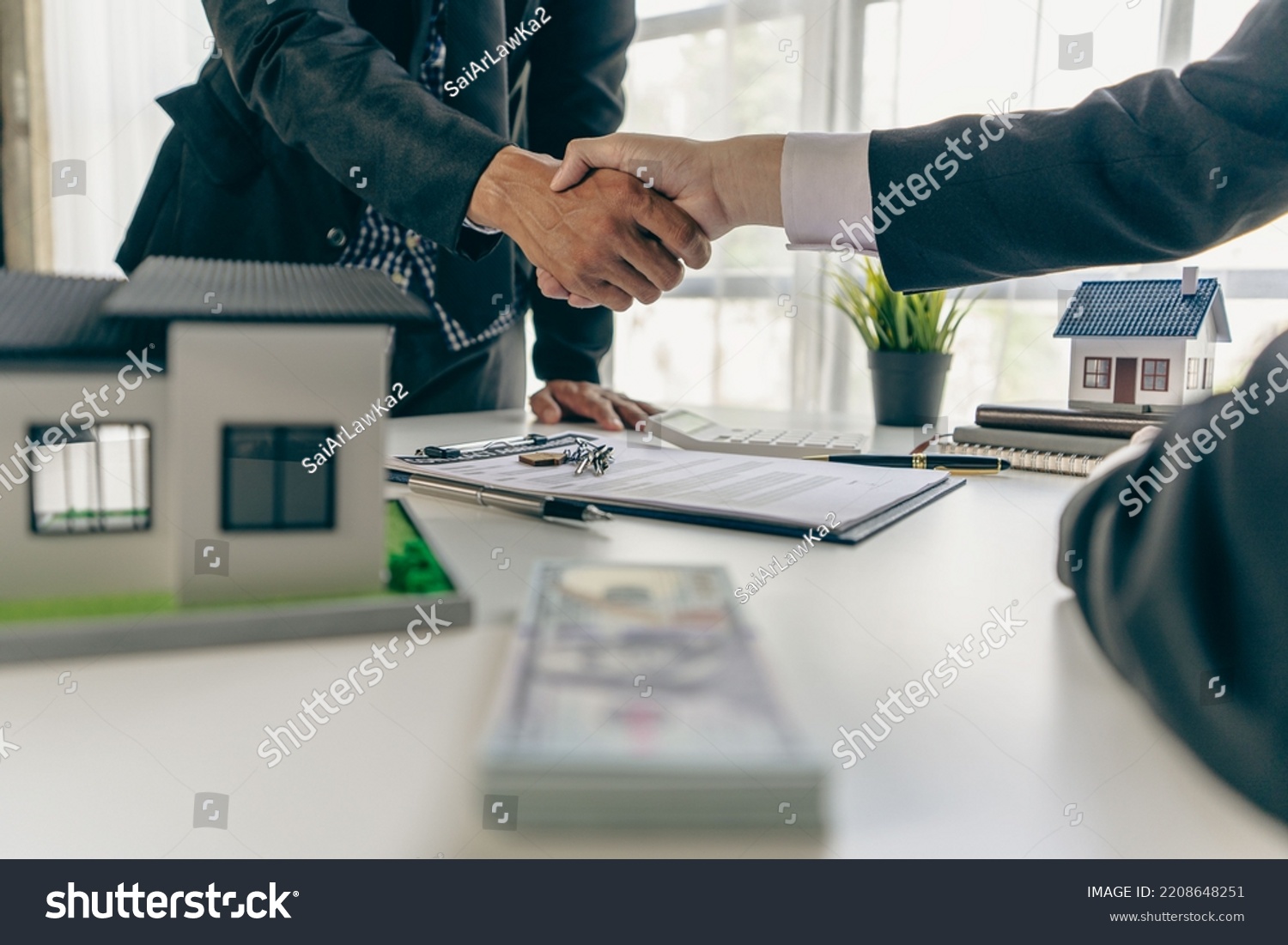 A real estate agent shakes hands with a client after completing a transaction regarding a house contract agreement. Contract documents and a house model on a wooden table. #2208648251
