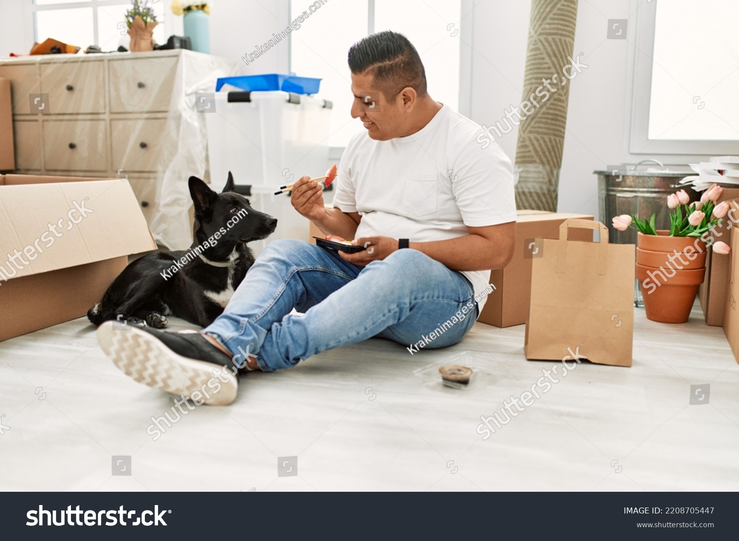Young latin man eating taka away sushi food sitting on the floor at new home with dog. #2208705447