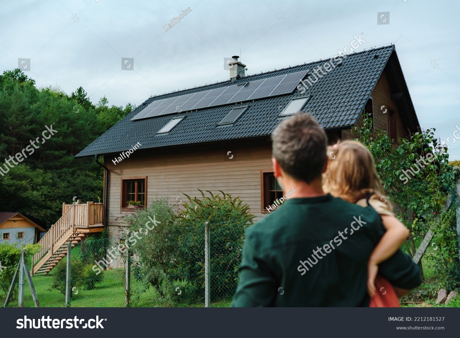 Rear view of dad holding her little girl in arms and looking at their house with installed solar panels. Alternative energy, saving resources and sustainable lifestyle concept. #2212181527