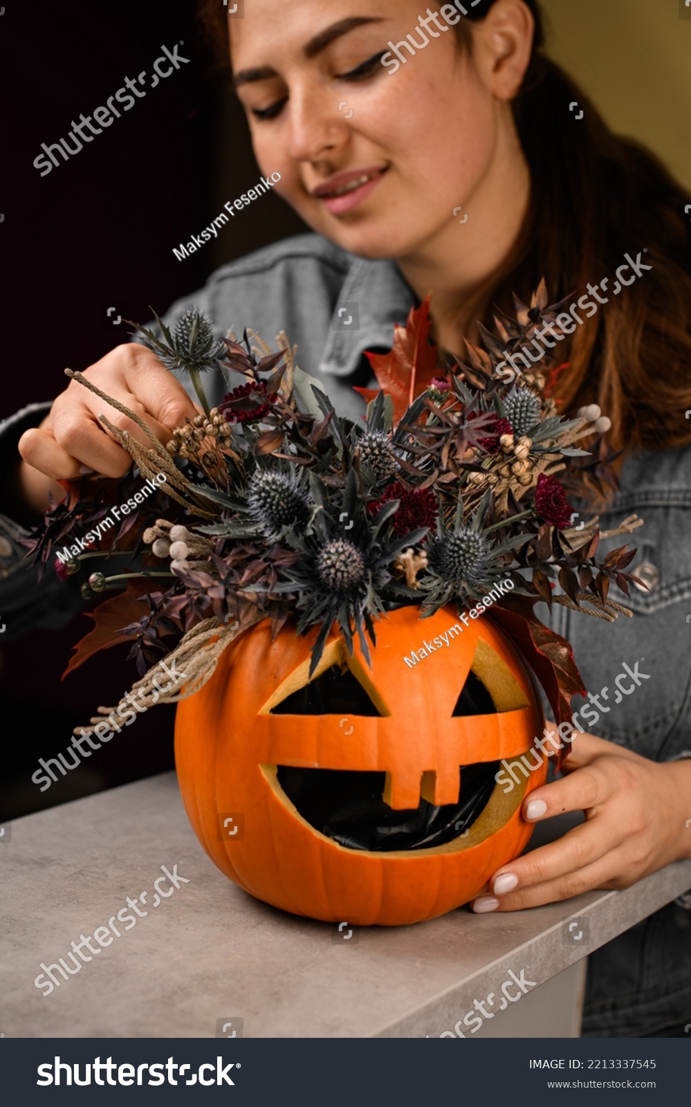 woman florist gently decorate lovely autumn arrangement of thistles flowers and plants in bright orange pumpkin with carved eyes and mouth. Beautiful floral decor for halloween. Close-up #2213337545