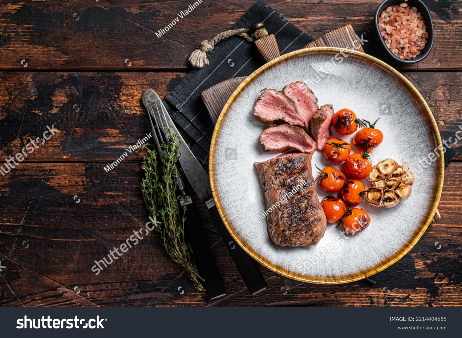 Roasted Lamb tenderloin meat in plate with grilled tomato and garlic, mutton sirloin fillet steak. Dark background. Top view. Copy space. #2214404585