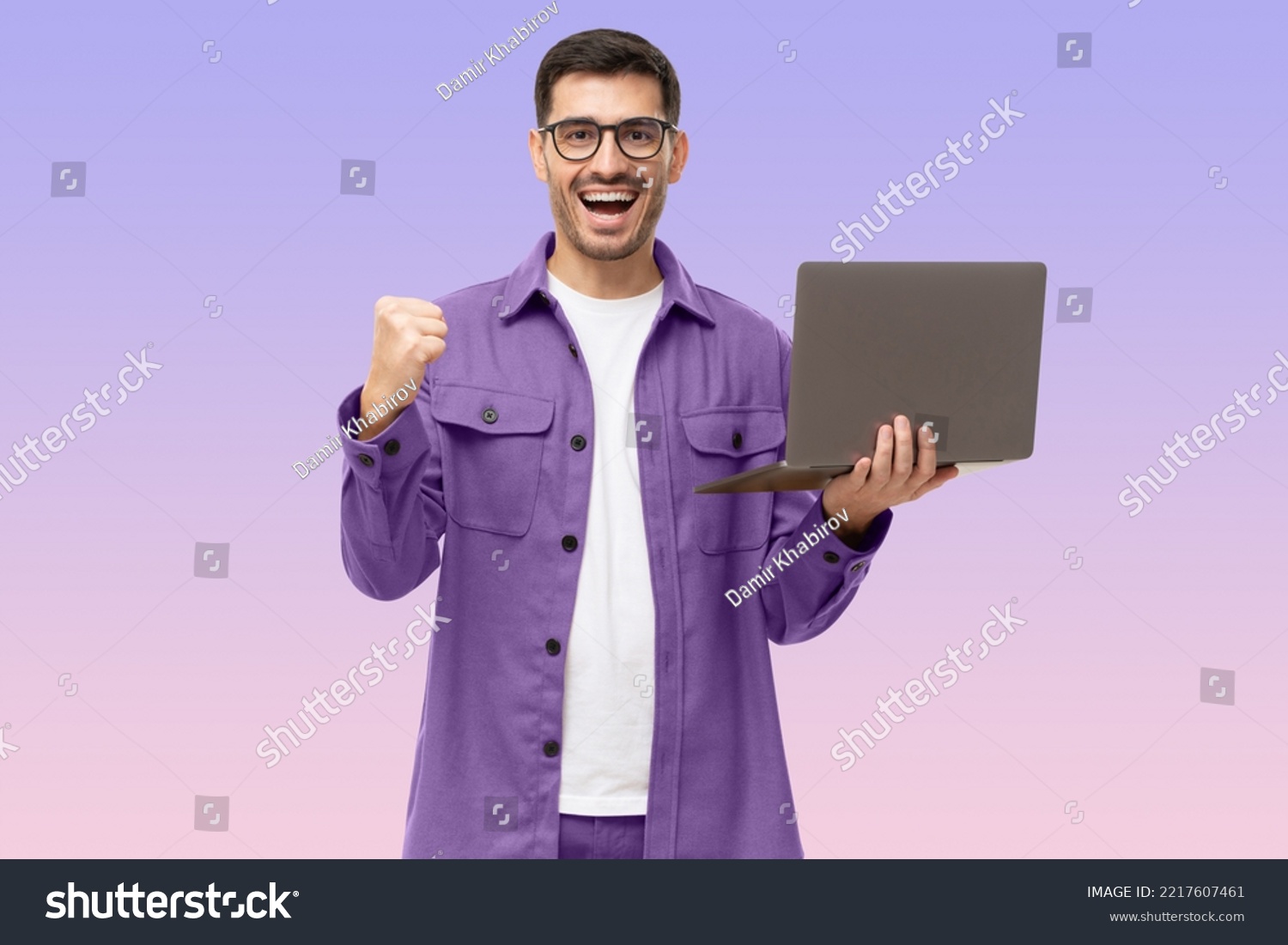 He's a winner! Happy young man in purple shirt looking at laptop screen with victory expression, isolated on purple background #2217607461