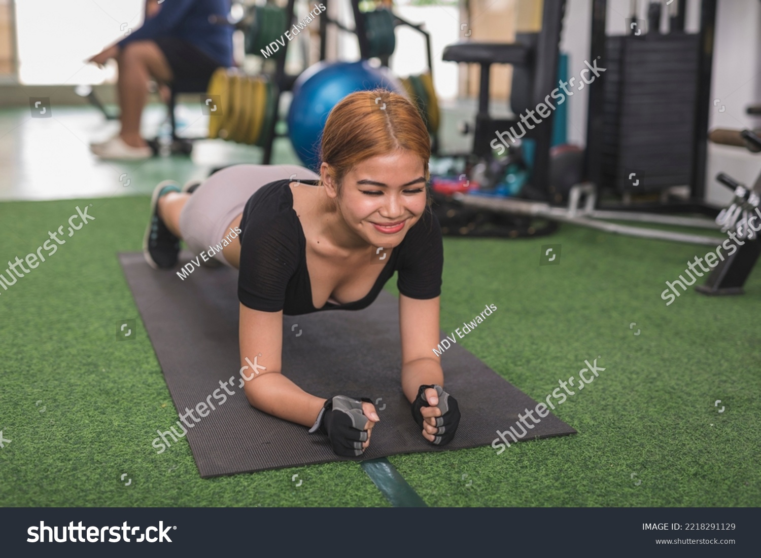 A charismatic woman smiles while doing planks on a black mat. Having fun and a lighthearted moment at the gym. #2218291129
