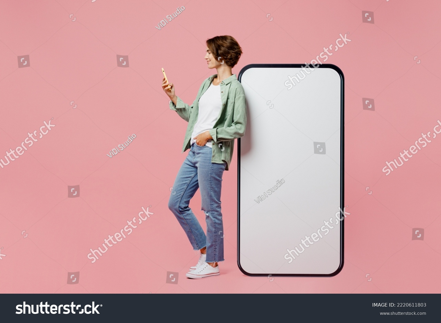 Full body side view young woman wear green shirt white t-shirt near big huge blank screen mobile cell phone smartphone with mockup use mobile cell phone isolated on plain pastel light pink background #2220611803