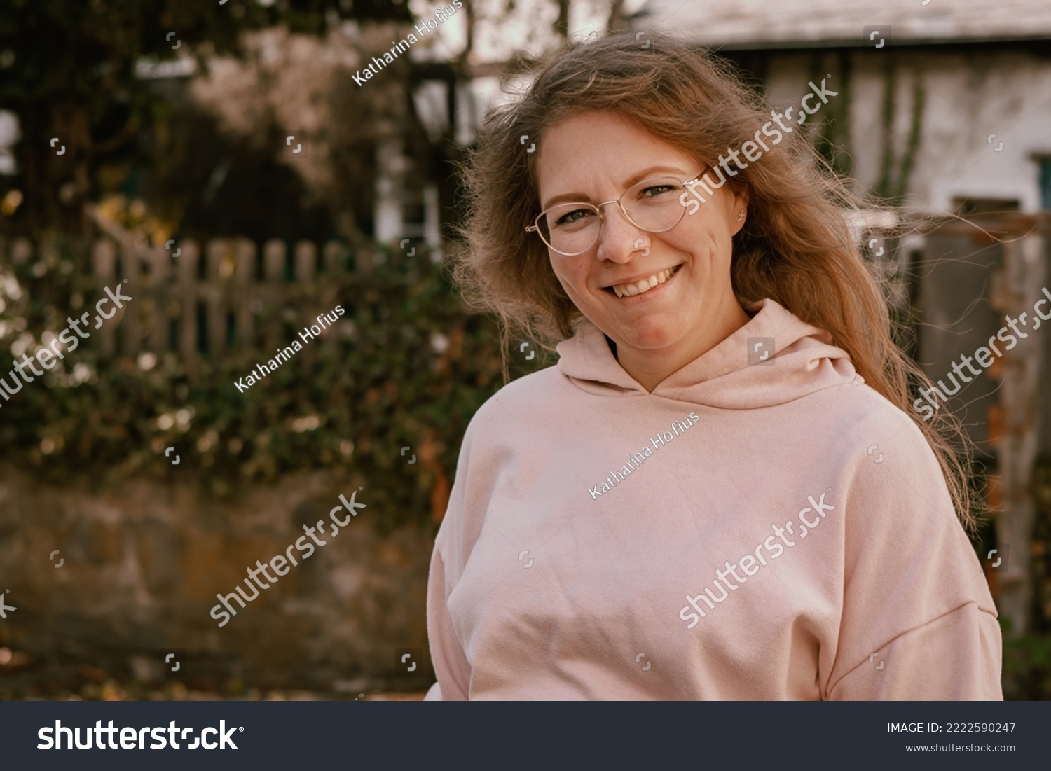 Close-up photography of a 30 years old woman showing a cute smile and looking straight into the camera; the woman is wearing a pink hoodie; the picture was taken outdoors in countryside #2222590247