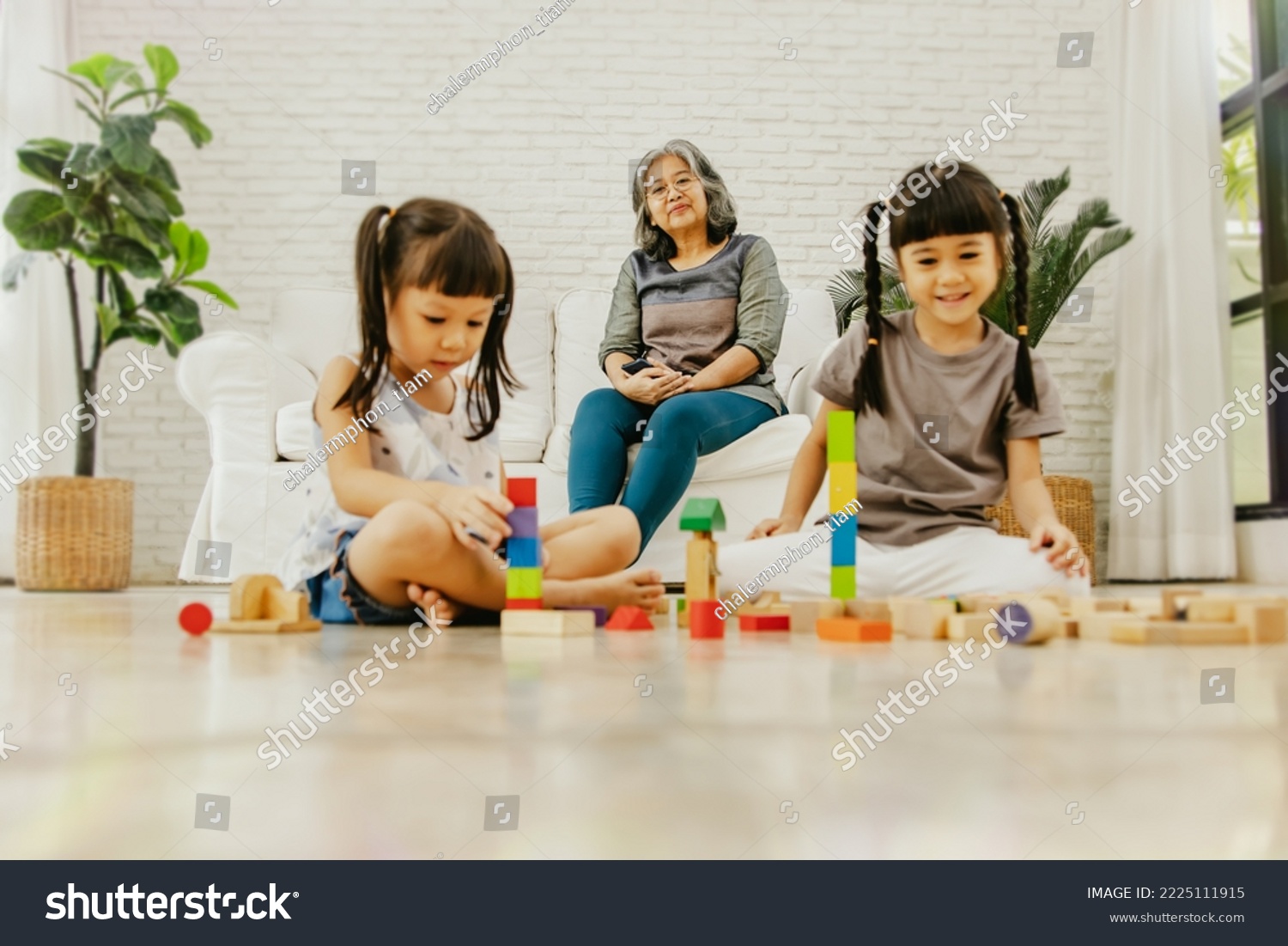Grandma sits on the sofa and watches two girls and granddaughters play with colorful wooden blocks that are non toxic to form tall tower with adorable ingenuity : Selected focus  #2225111915