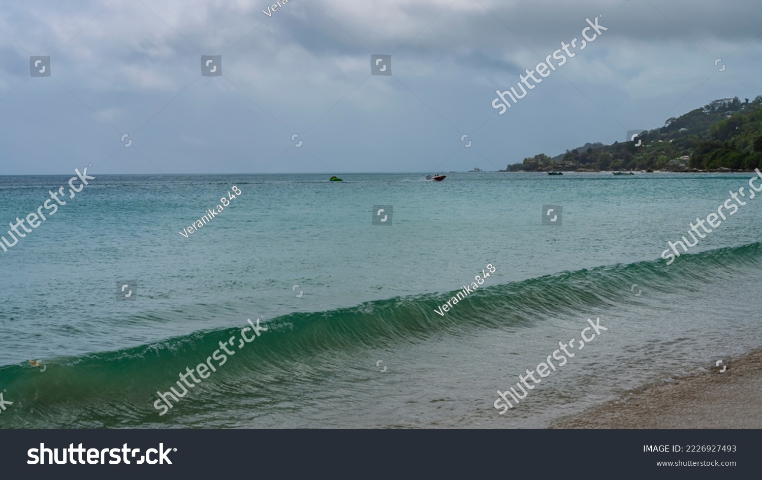 A long turquoise wave rolls onto the sandy shore. Boats are visible on the surface of the ocean. A green hill against a cloudy sky. Seychelles. Mahe. Beau Vallon beach #2226927493