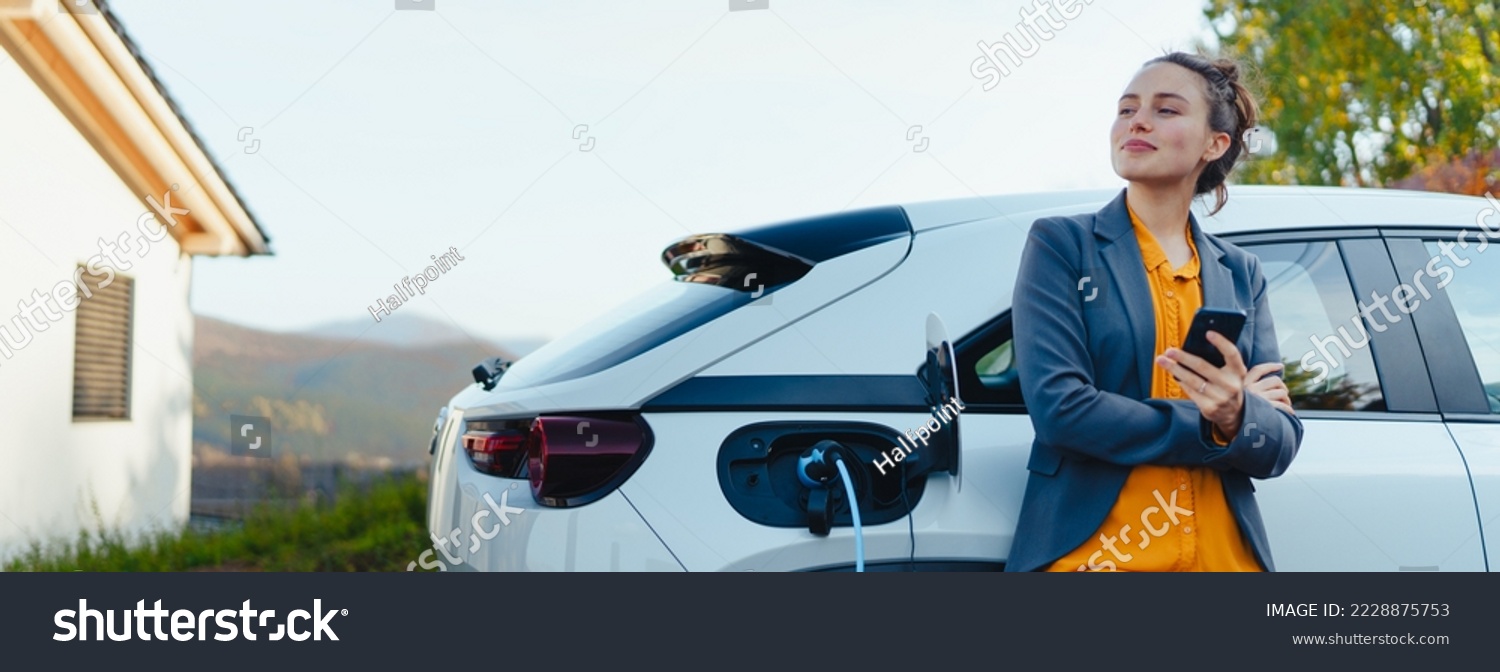 Young woman with smartphone waiting while her electric car charging in home charging station, sustainable and economic transportation concept. #2228875753