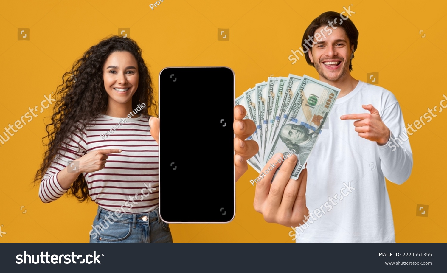 Cheerful young lovers man and woman gambling on Internet, using cell phone with black empty screen, pointing at smartphone, bunch of dollars, yellow studio background, mockup. Online bet concept #2229551355