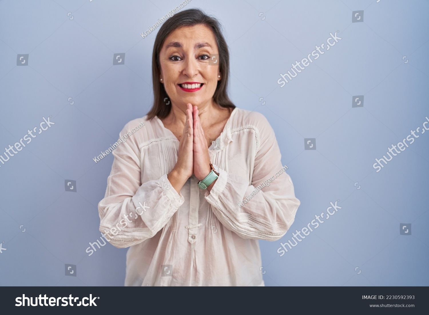 Middle age hispanic woman standing over blue background praying with hands together asking for forgiveness smiling confident.  #2230592393