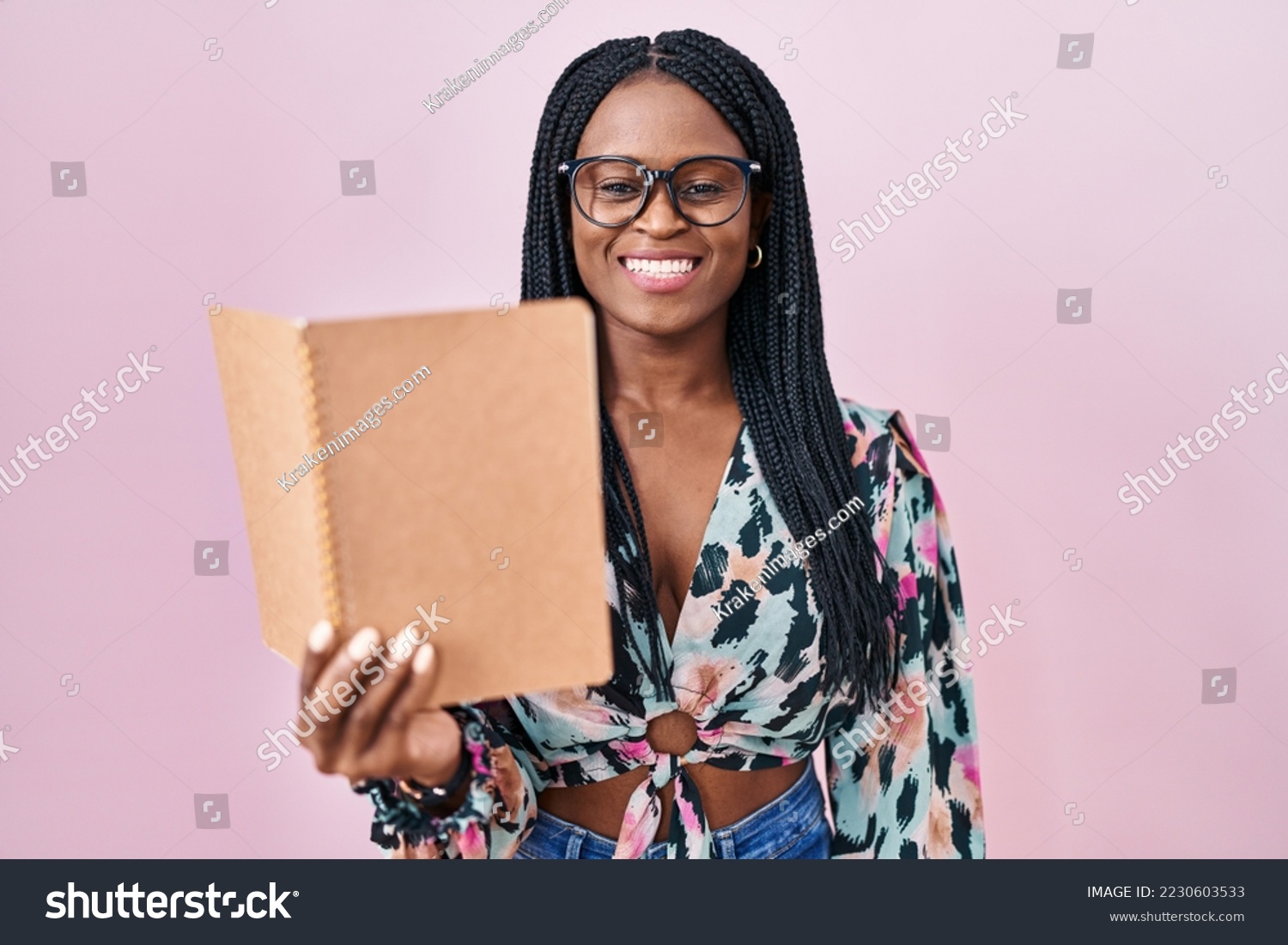 African woman with braids reading a book looking positive and happy standing and smiling with a confident smile showing teeth  #2230603533