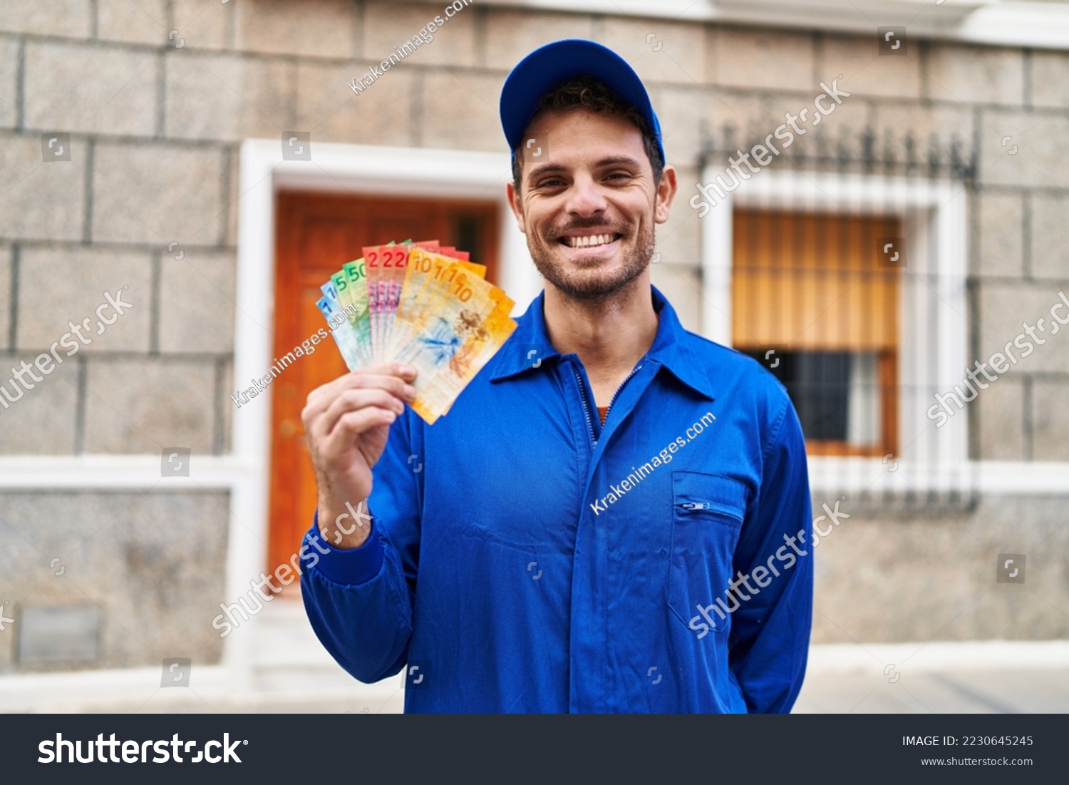 Young hispanic man wearing handyman uniform holding swiss francs looking positive and happy standing and smiling with a confident smile showing teeth  #2230645245